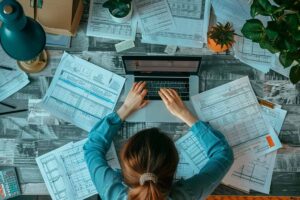 Person working on a laptop with financial documents spread out on a desk, appearing overwhelmed by financial planning and investment strategies.